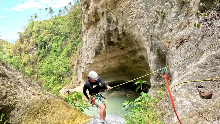 Guest on rope above a cave pool, with palm trees on the canyon rim